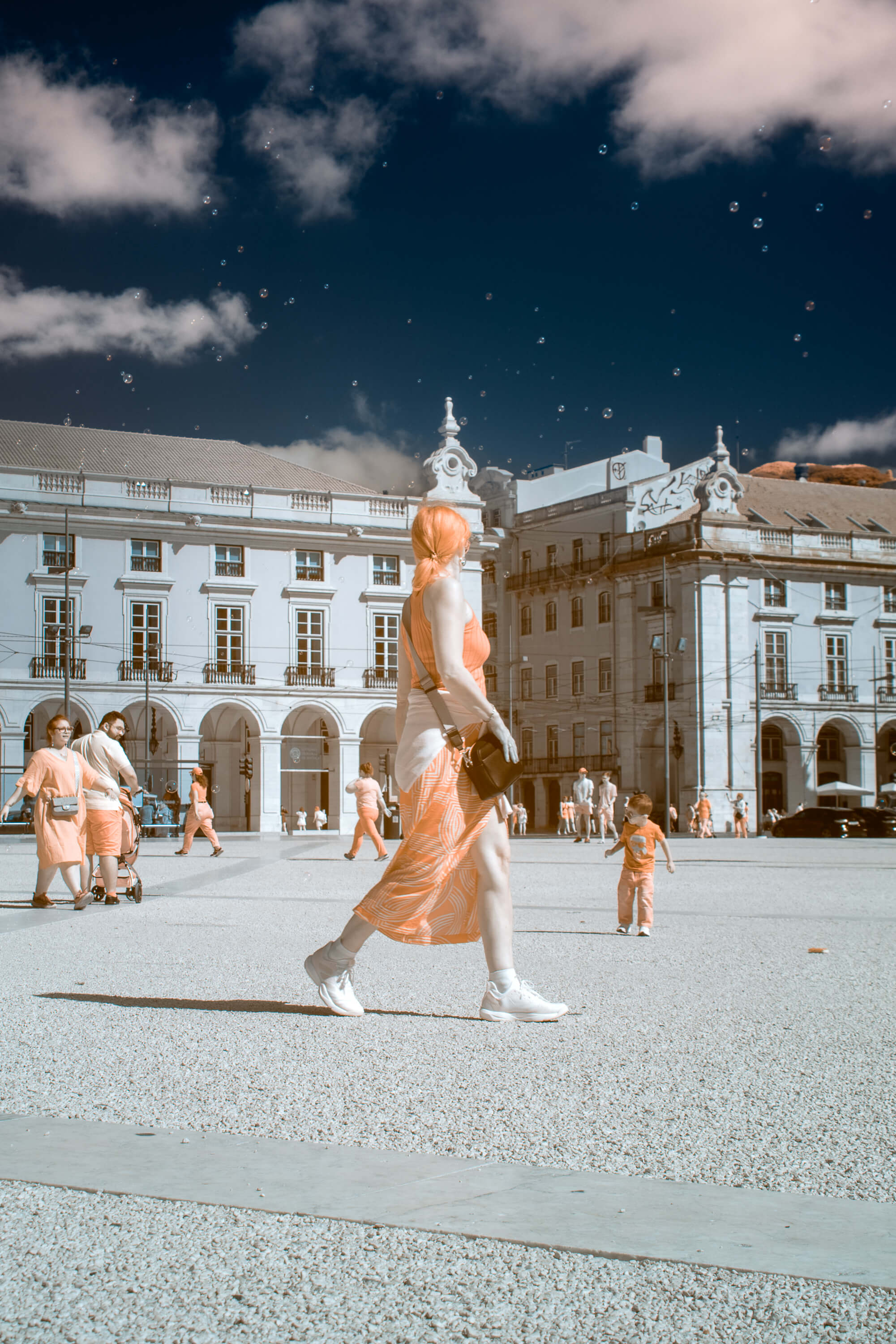 A woman walks across Praça do Comércio while staring at bubbles floating by. Captured by an infrared sensitive camera that creates unnatural colors.