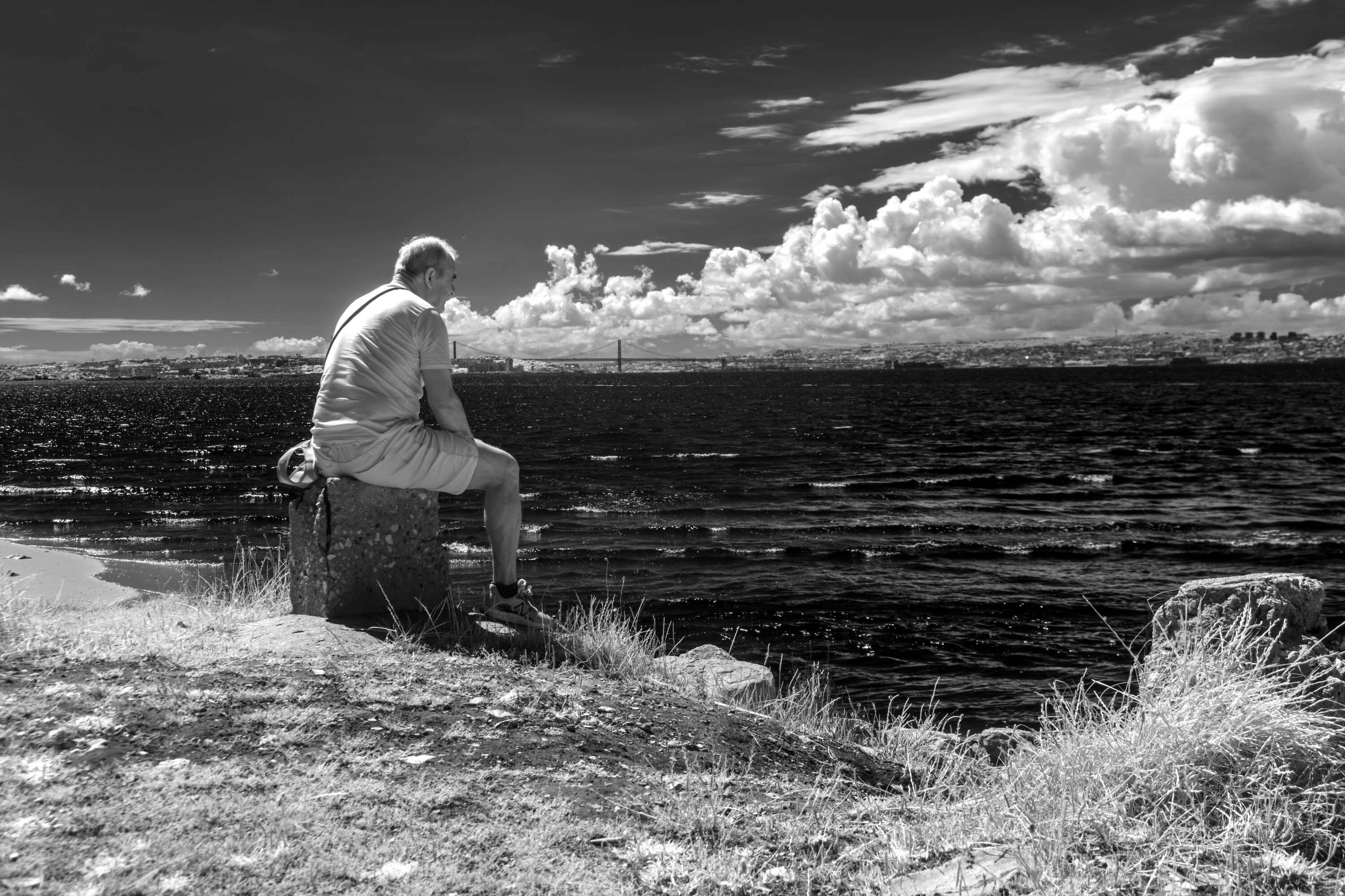 An older man in Barreiro sits by the Tagus staring outward. The clouds behind him almost appear like he is breathing them out.