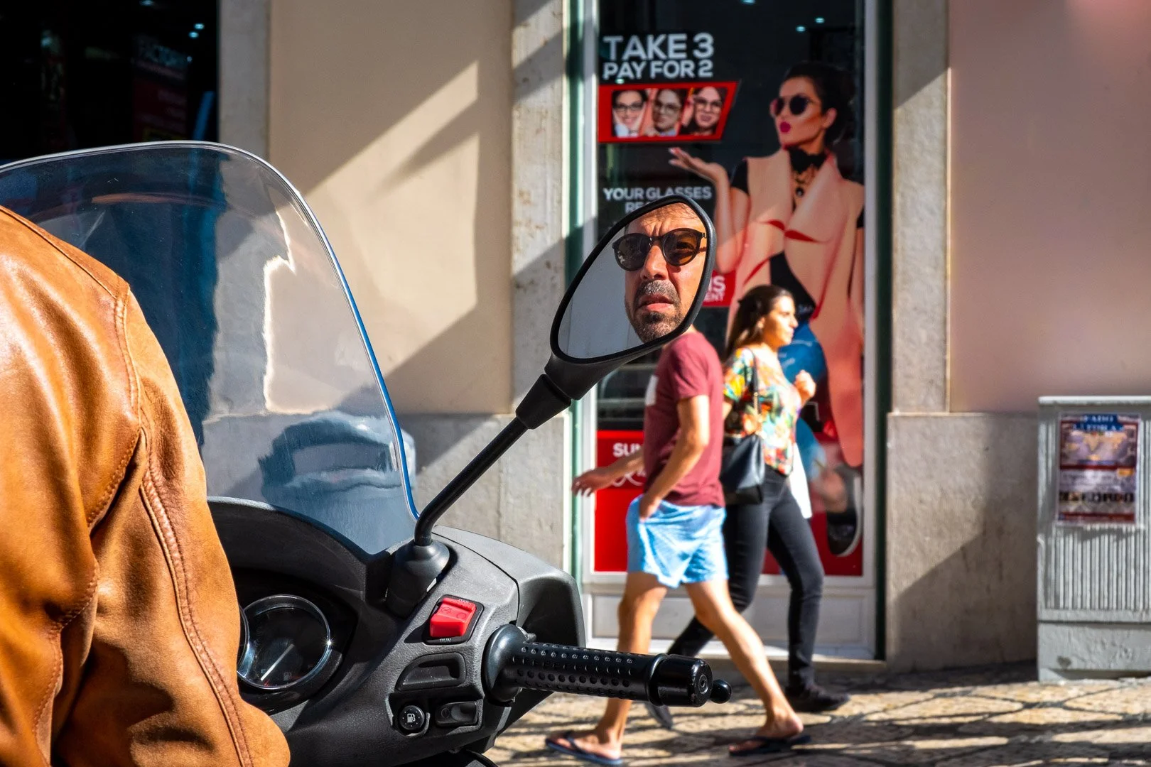 A man's face, reflected in a motorbike wing mirror, stares out from a busy Lisbon street, an optician's advertisement visible behind. Photograph by Gil Ribeiro.