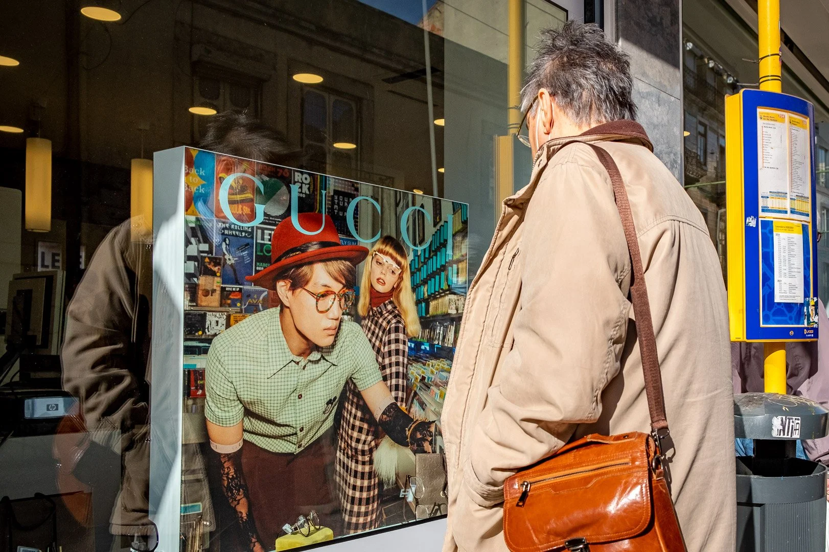 An older man with a brown leather bag examines a Gucci window display on a sunlit city street. Photograph by Gil Ribeiro.