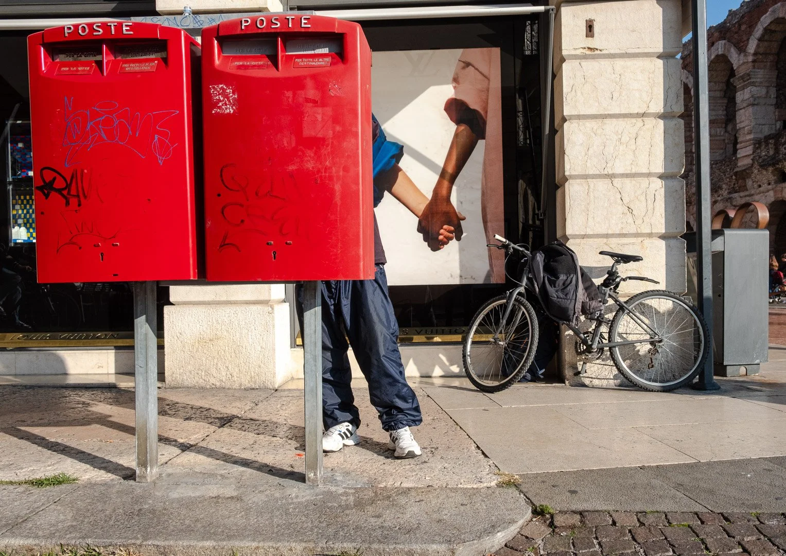 A person stands obscured behind two red Italian post boxes, a Louis Vuitton advertisement showing clasped hands visible behind them. Photograph by Gil Ribeiro.