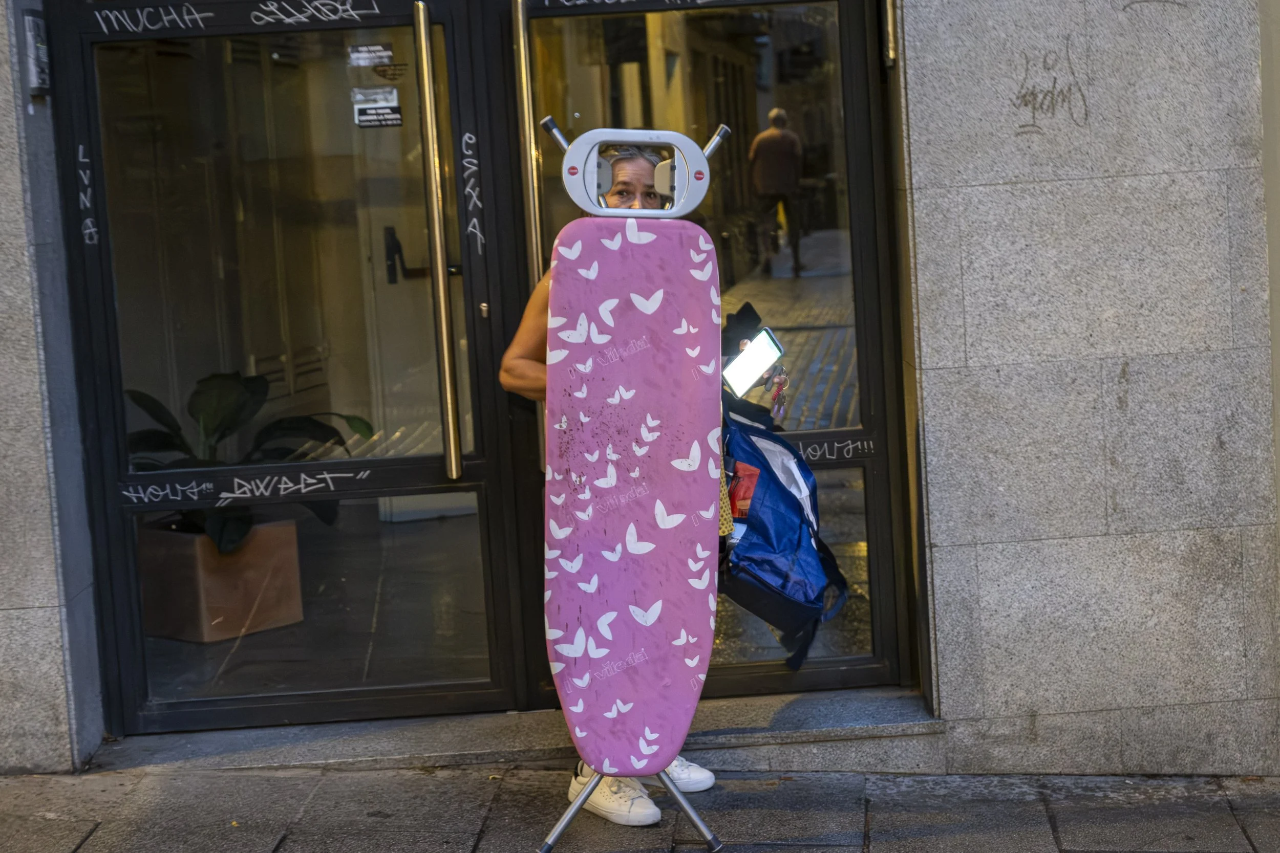 A woman carries a large pink ironing board vertically in front of her on a city street, her face visible through the board's small mirror. Photograph by Gil Ribeiro.