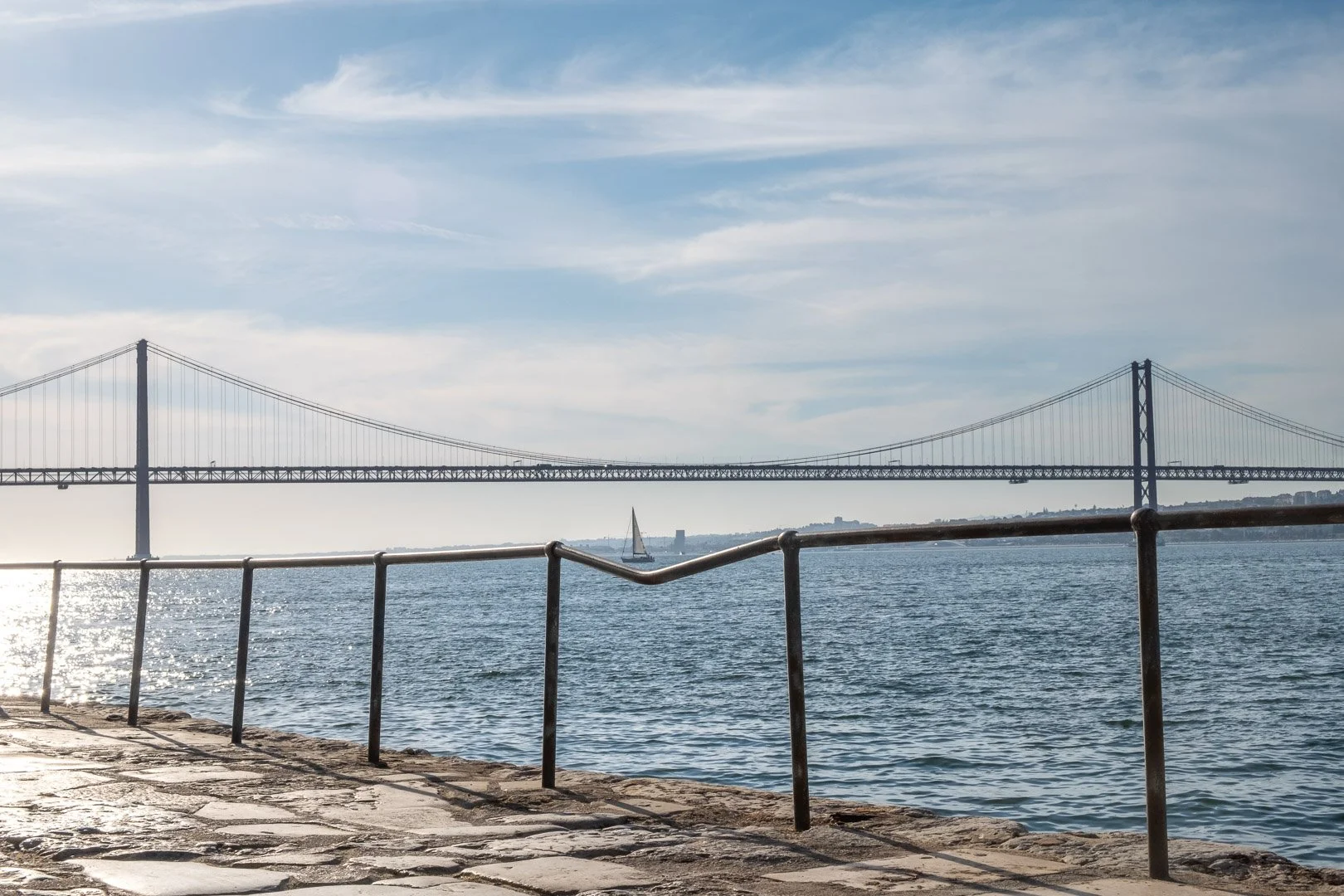 A sailboat crosses the Tagus beneath the Ponte 25 de Abril, framed between iron railings on a cobbled riverside path. Photograph by Gil Ribeiro.