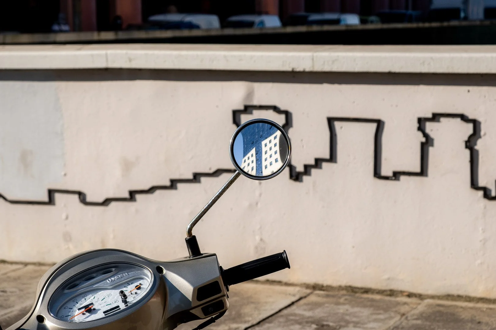 A scooter's wing mirror reflects a high-rise building, set against a wall painted with a pixelated city skyline. Photograph by Gil Ribeiro.