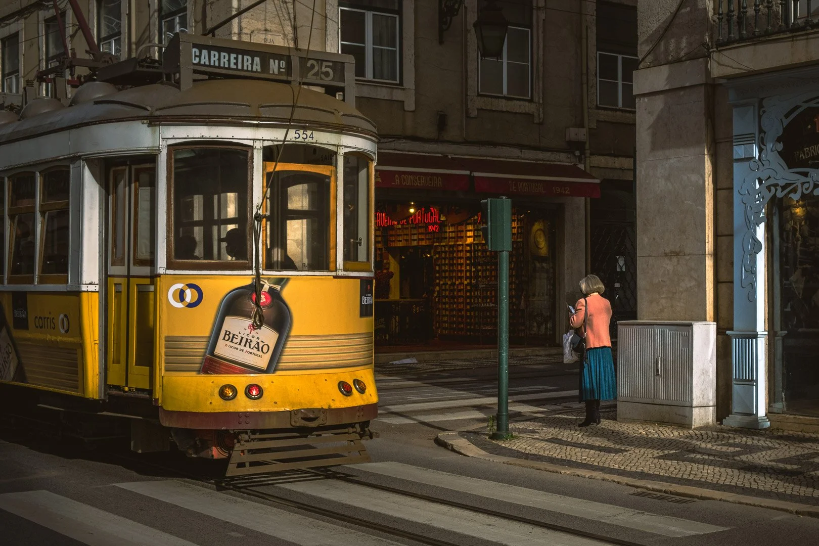Lisbon's classic yellow tram rounds a corner in warm evening light as a woman in an orange jacket waits at the crossing. Photograph by Gil Ribeiro.