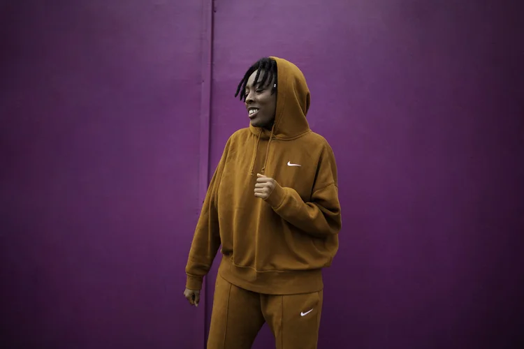 A person in a brown Nike hoodie stands laughing against a vivid purple wall. Photograph by Will Bremridge.