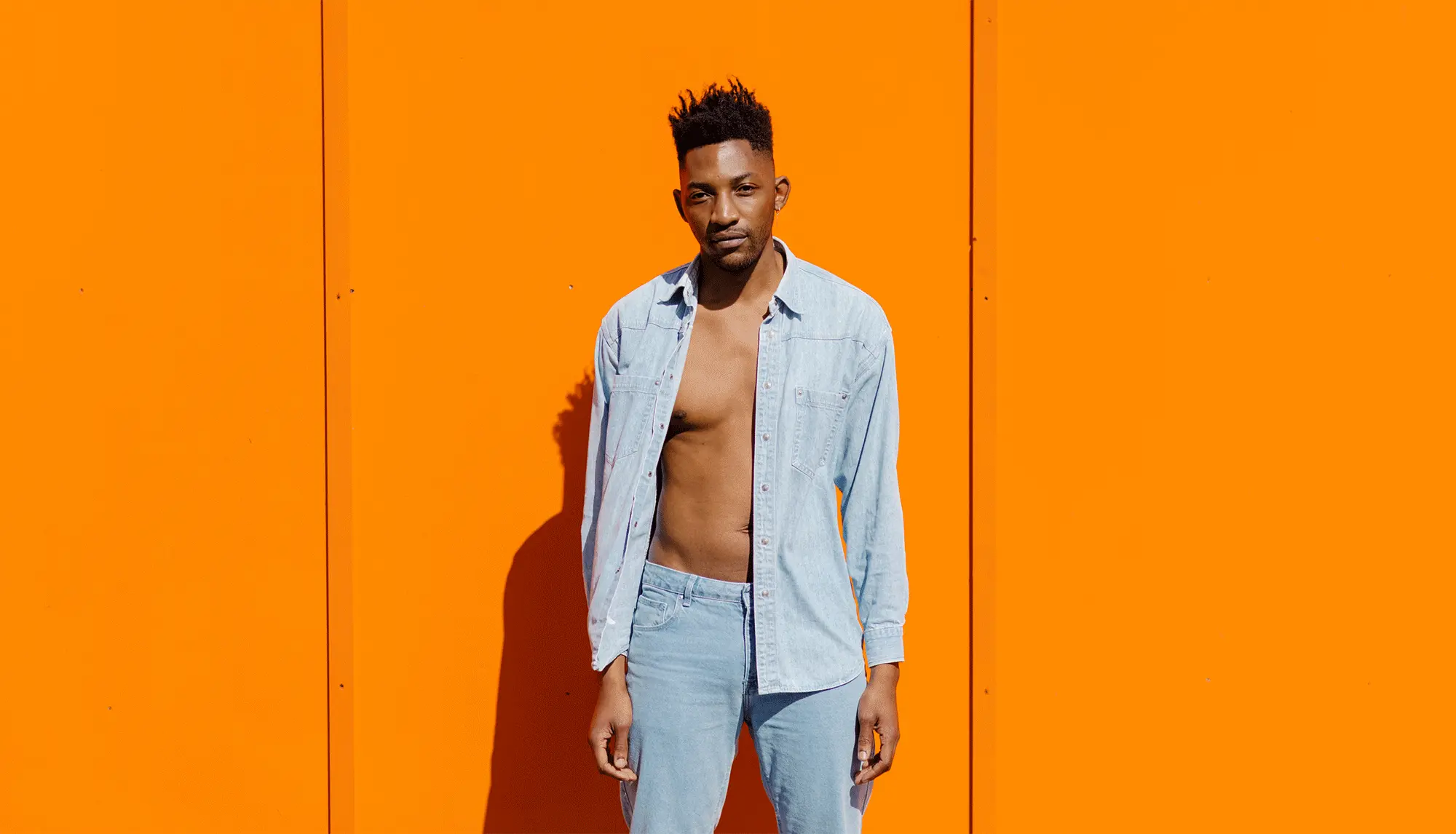 A man in an open denim shirt stands against a bold orange wall in bright sunlight. Photograph by Will Bremridge.