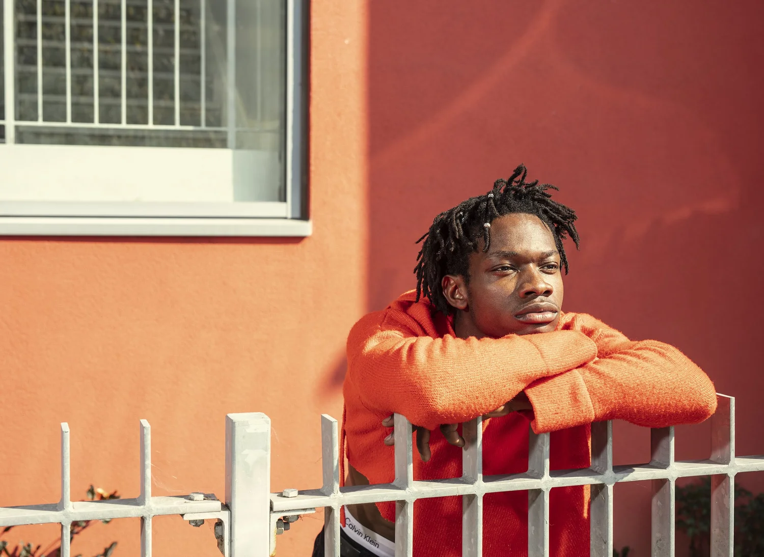 A young man in an orange hoodie rests his arms on a white iron fence against a deep red wall. Photograph by Will Bremridge.
