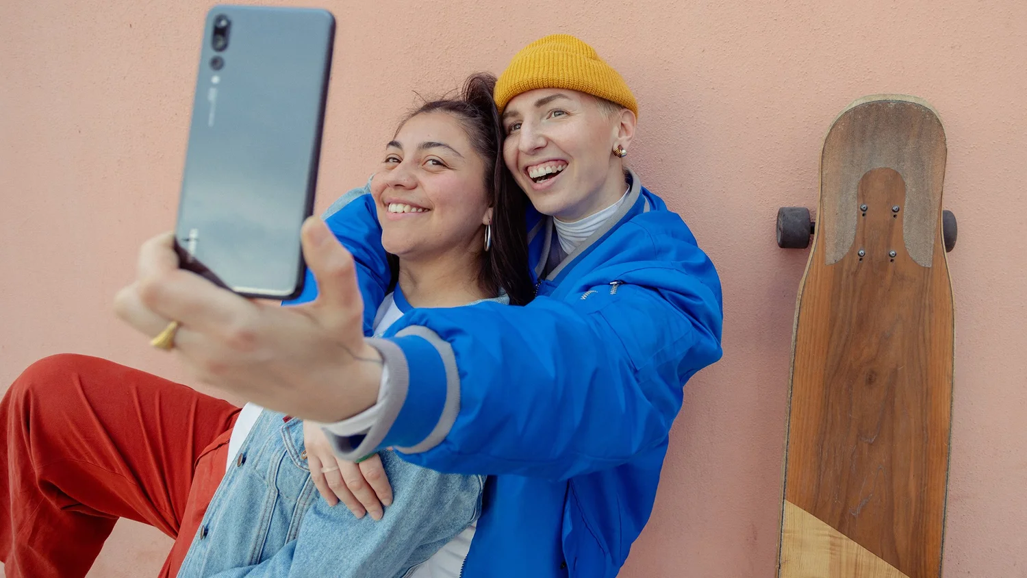 Two women laugh and take a selfie against a pink wall. Photograph by Will Bremridge.