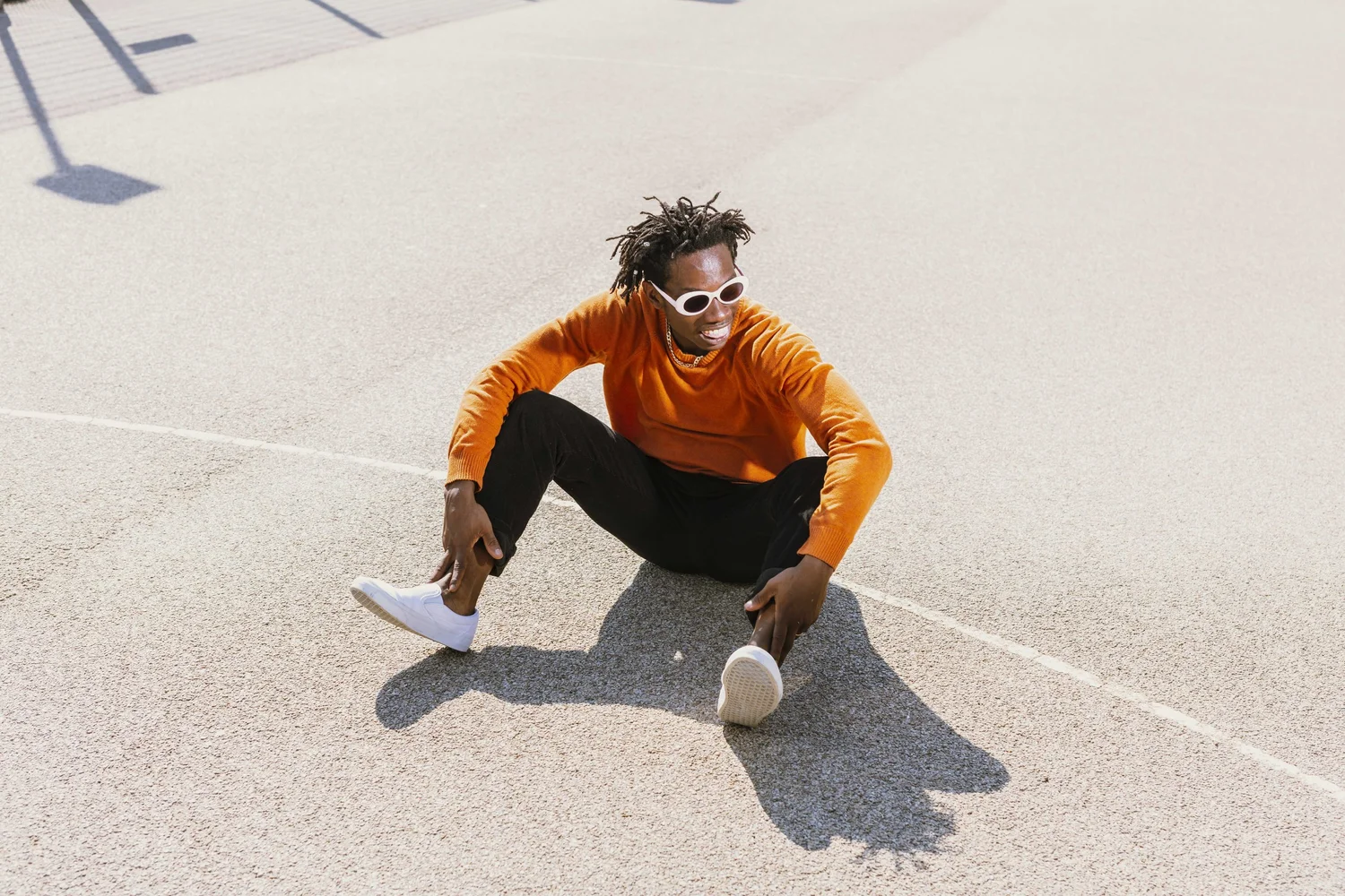 A young man in an orange jumper sits on a grey outdoor court wearing white oval sunglasses, smiling. Photograph by Will Bremridge.