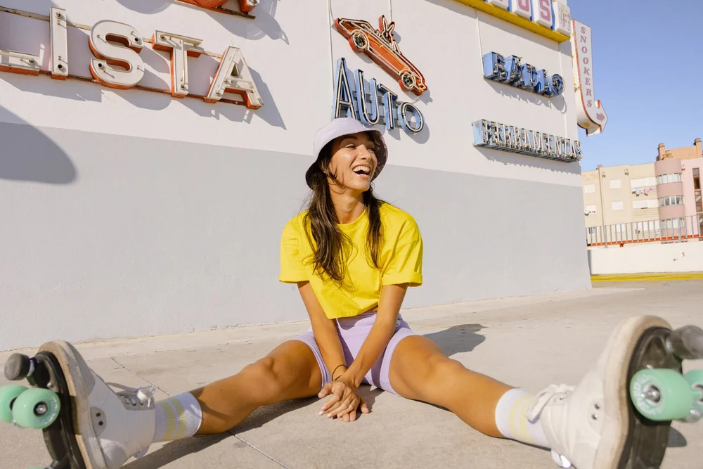 A young woman in a yellow t-shirt and lavender shorts sits laughing wearing roller skates. Photograph by Will Bremridge.