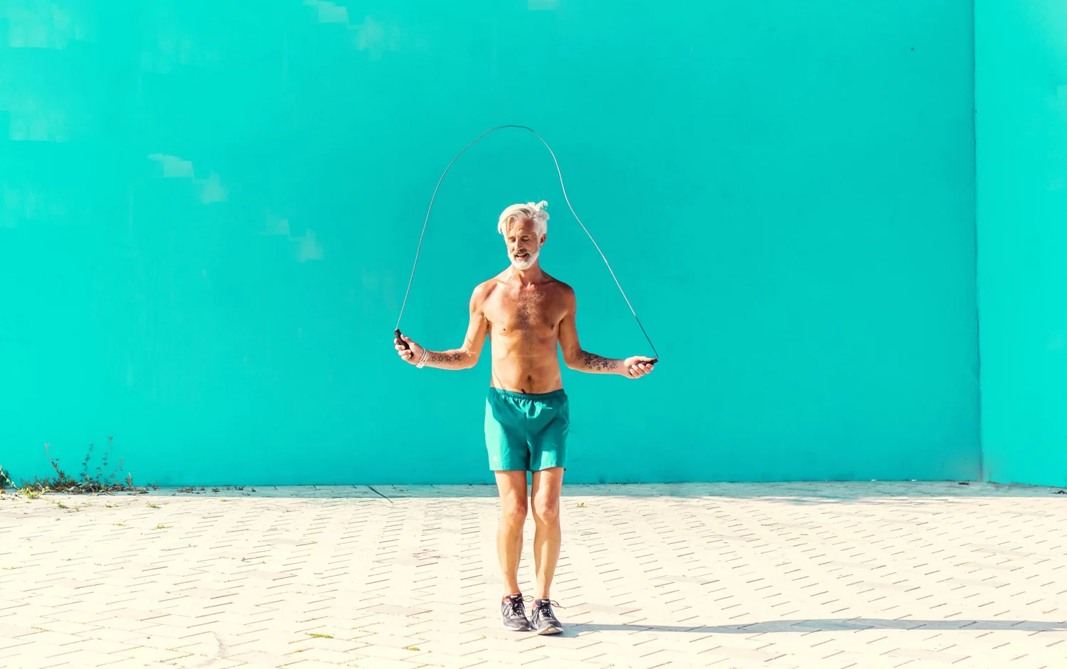 An older man with white hair skips rope shirtless against a vivid teal wall. Photograph by Will Bremridge.