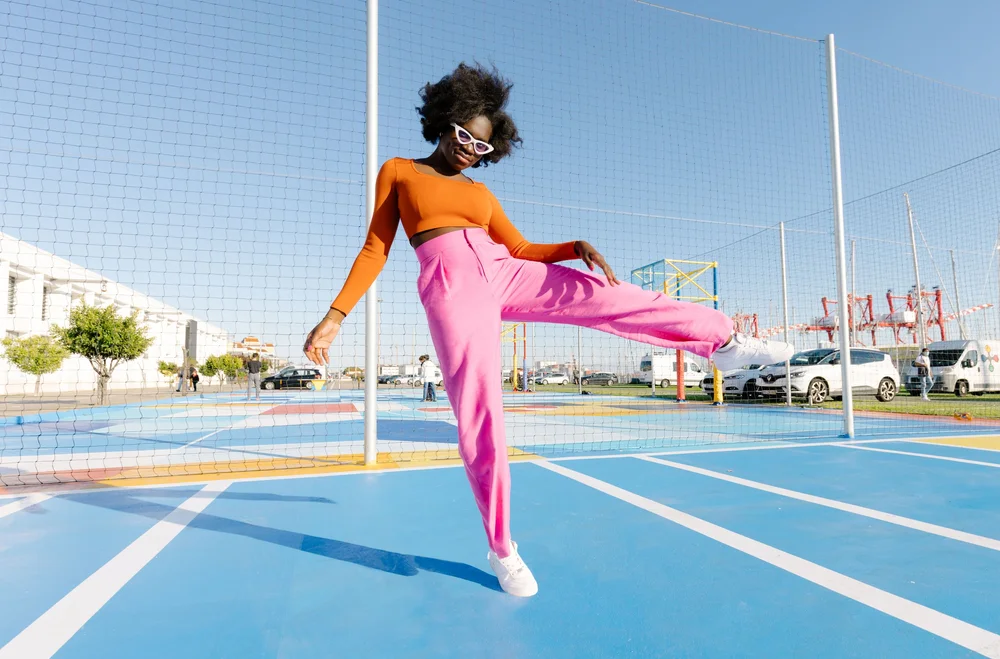 A woman in an orange crop top and pink trousers kicks her leg high on a blue outdoor sports court. Photograph by Will Bremridge.