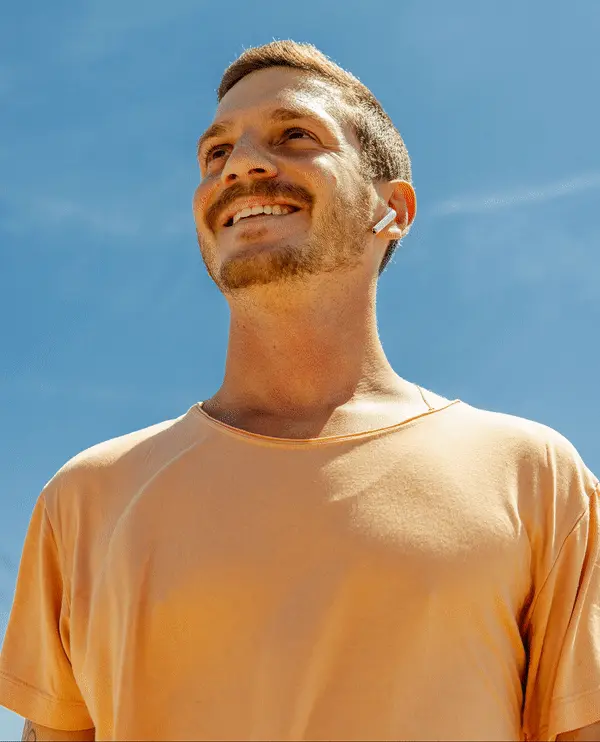 A smiling man in a peach t-shirt with earbuds looks up at a clear blue sky. Photograph by Will Bremridge.